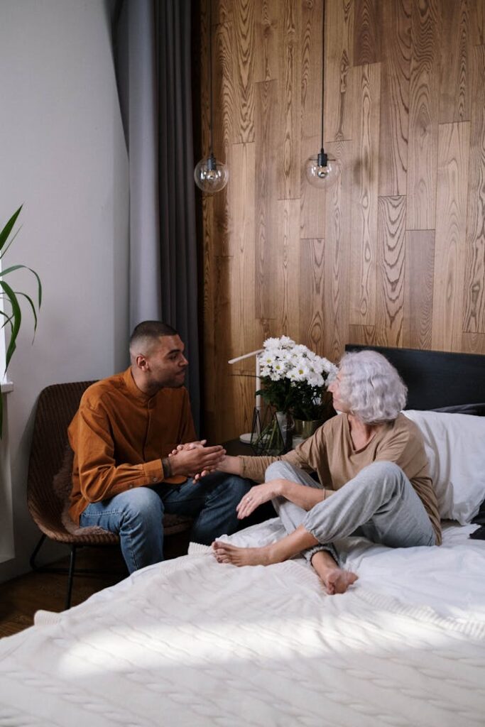 A young man and elderly woman have a heartfelt conversation in a cozy bedroom setting.